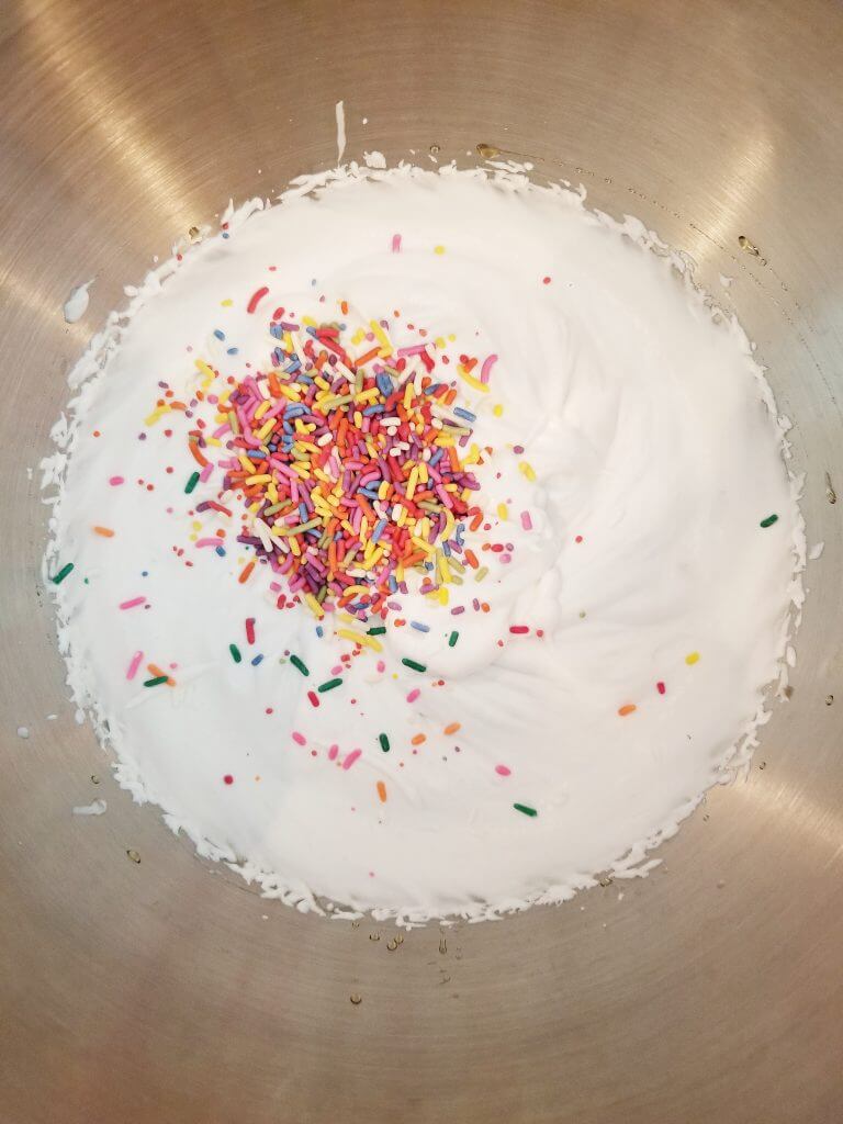 rainbow sprinkles on top of whipped coconut cream in a stainless steel mixing bowl
