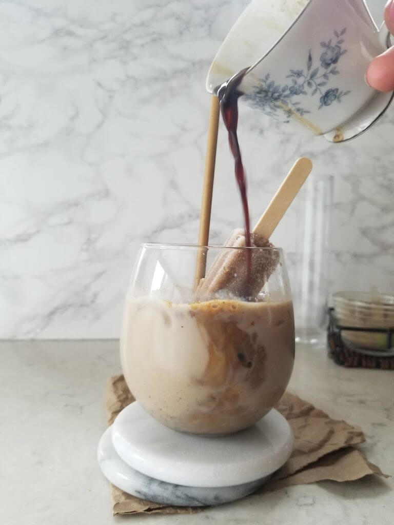 pouring a shot of espresso from a white and blue teacup into the stemless wine glass with the iced matcha maca cacao