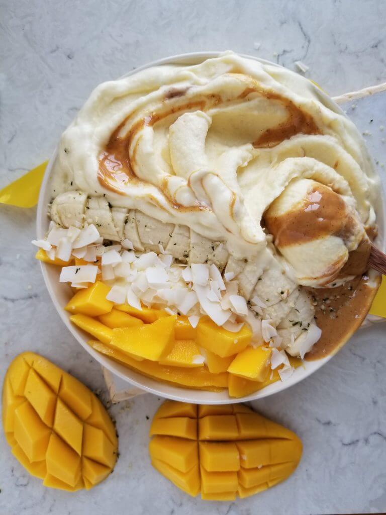 pineapple banana smoothie bowl topped with banana, coconut chips, mango, hemp seeds with peanut butter swirled in