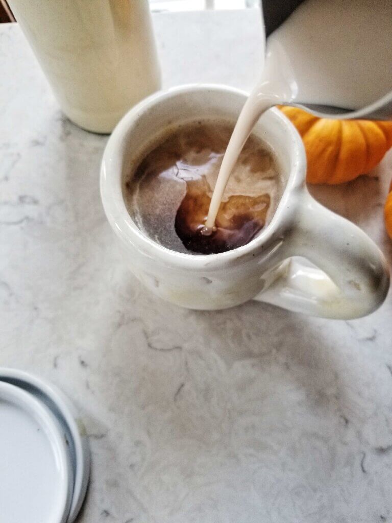 pouring almond milk pumpkin spice creamer into coffee in a white mug